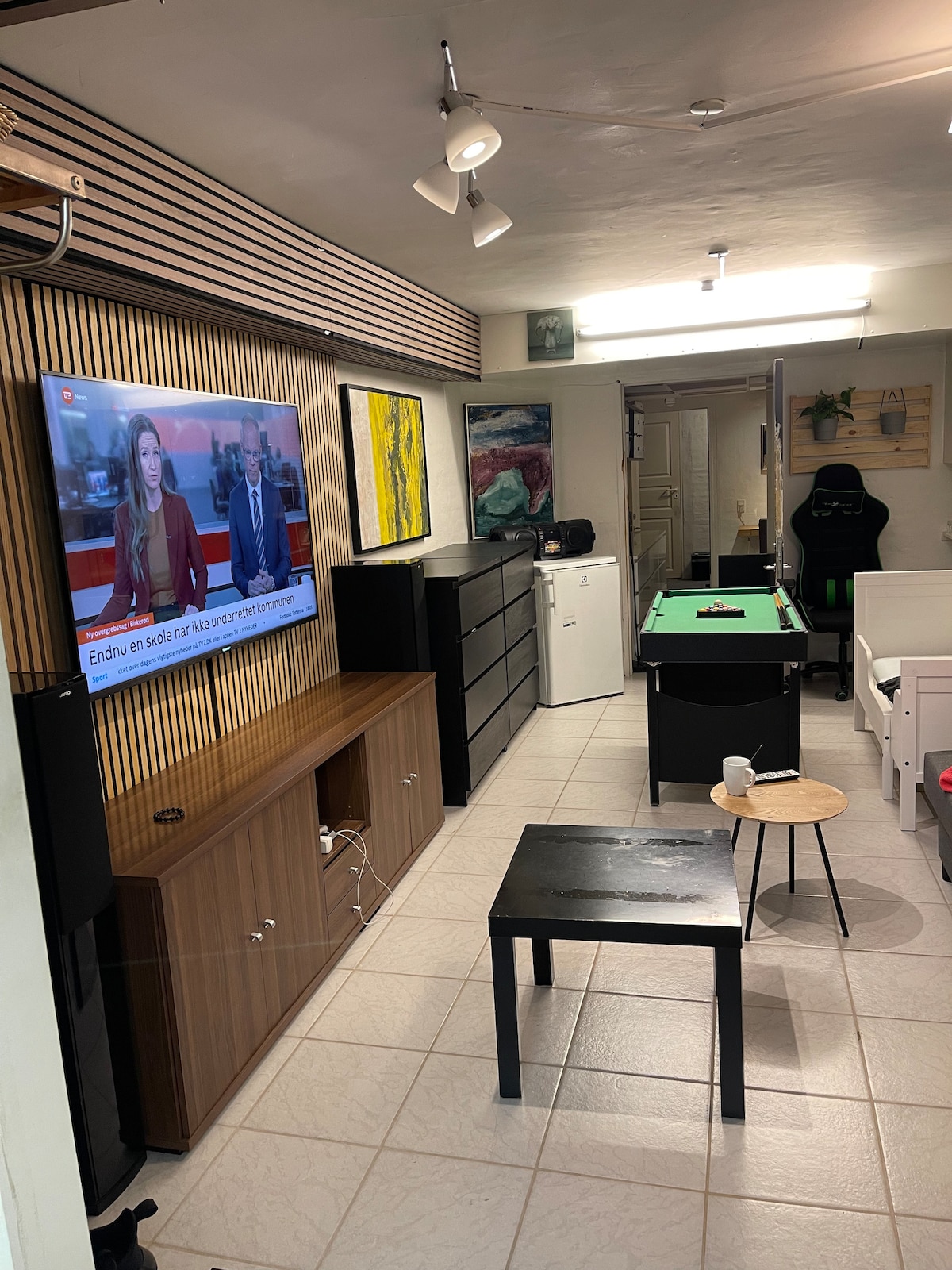 A cozy living area is shown, featuring a large television mounted on a wooden panel. A green pool table occupies the space, complemented by a small round table and a black rectangular table. A white couch and a refrigerator are visible in the background.