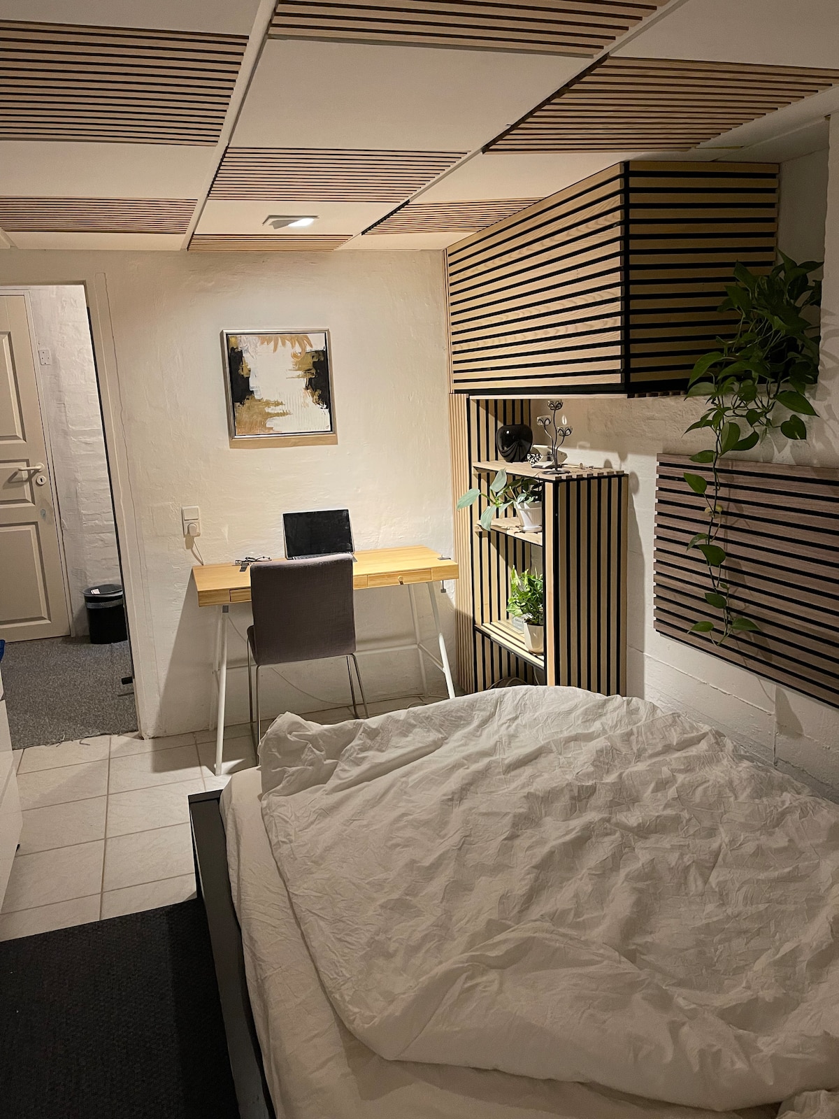 A compact room features a spacious bed with a white comforter and simple bedding. A small desk with a chair is positioned near a wall-mounted shelf adorned with greenery. Soft lighting complements the wooden accents throughout the space, offering a calming ambiance.