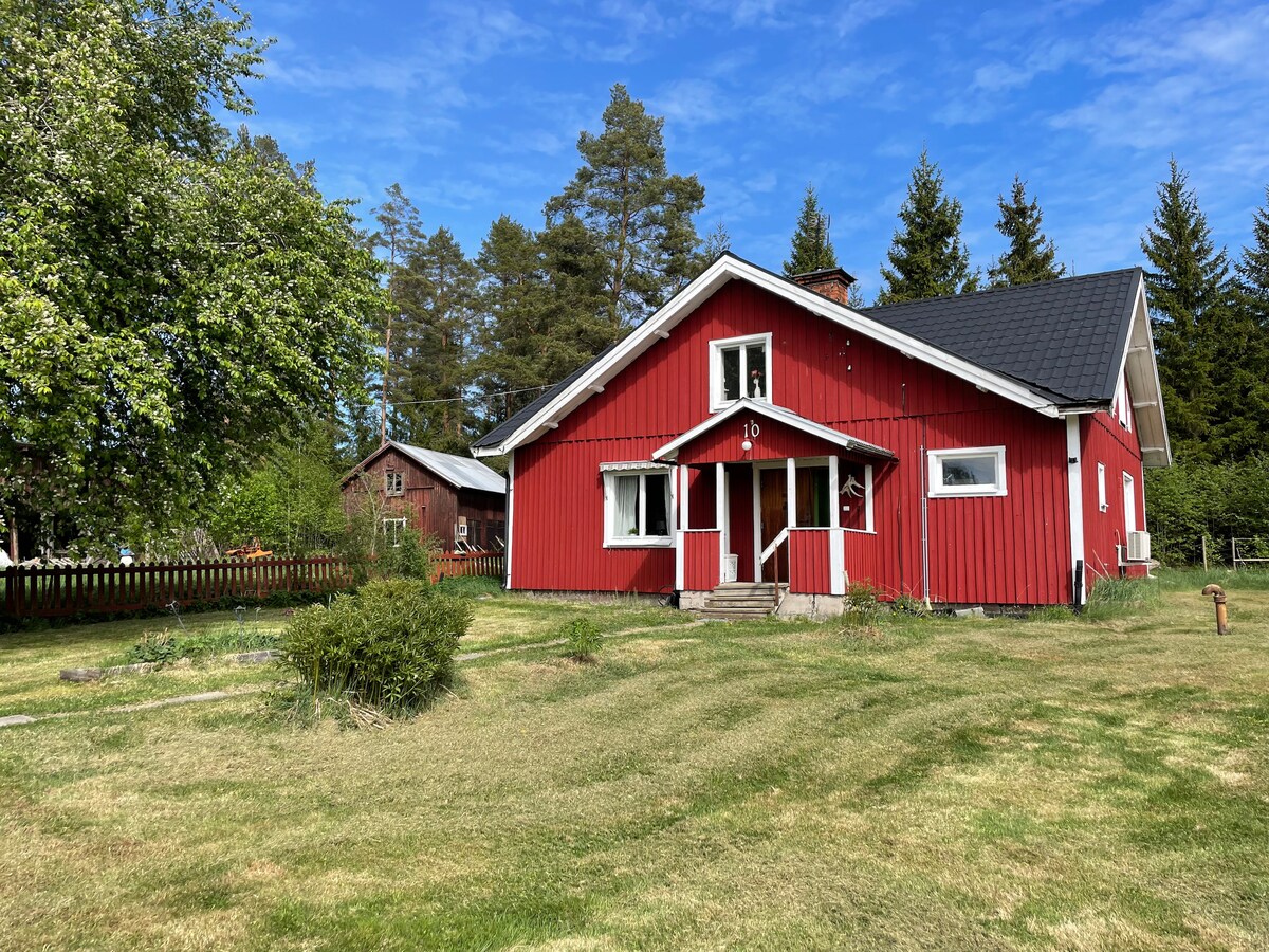 A red house with white trim is set in a spacious green yard, surrounded by trees. The front features a welcoming entrance with steps leading up. The roof is covered with dark shingles, and the sky above is bright and clear.