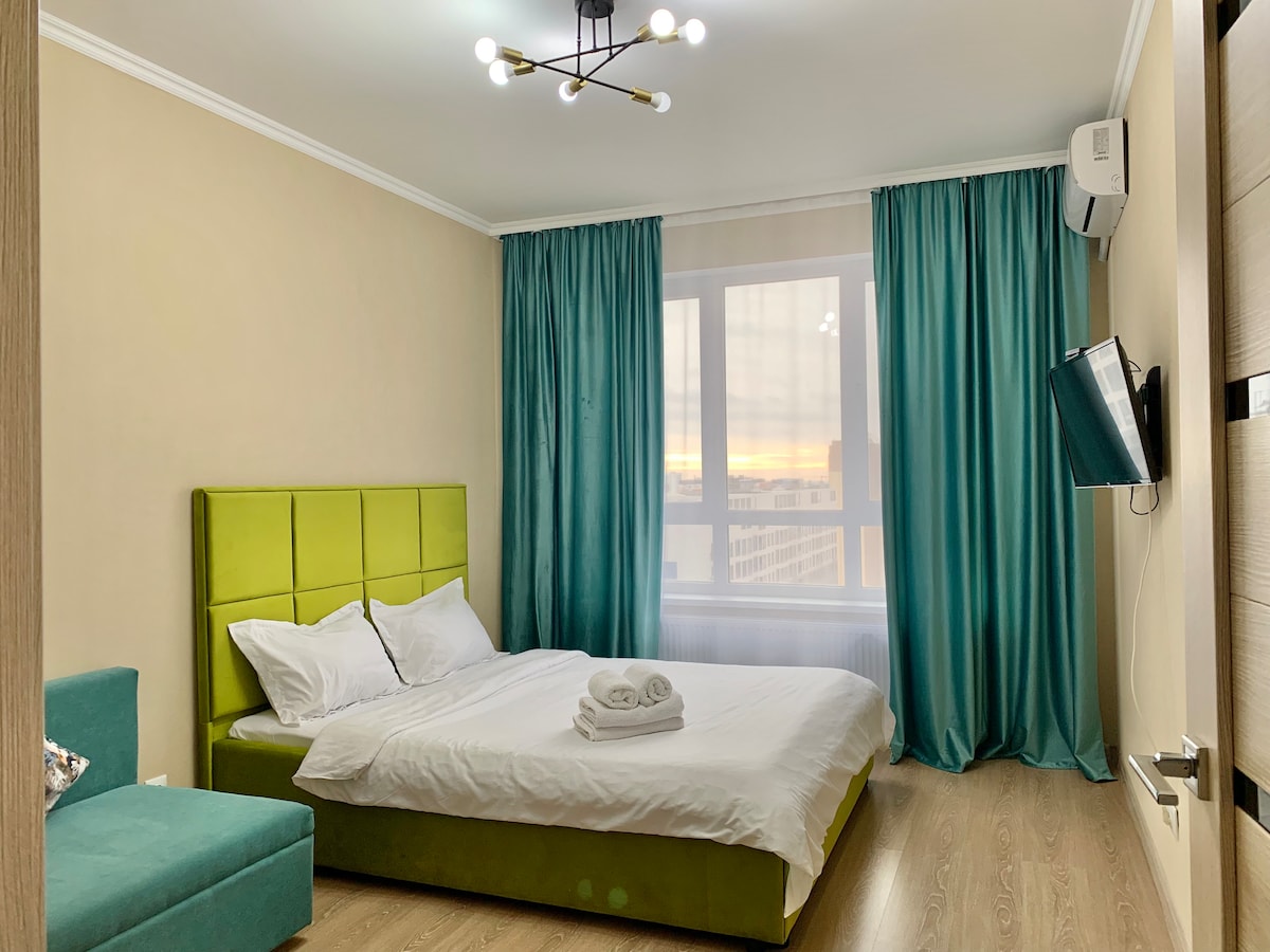 A bright bedroom features a spacious bed with white linens, a green headboard, and a cozy armchair. Floor-to-ceiling windows draped in teal curtains offer a view of the horizon, while a wall-mounted television is visible across the room.