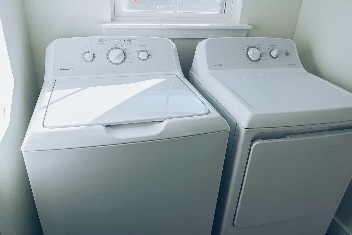 A white washing machine and dryer are positioned next to each other, featuring simple control dials on the front. Light streams in from a nearby window, illuminating the clean and functional design of the laundry appliances.