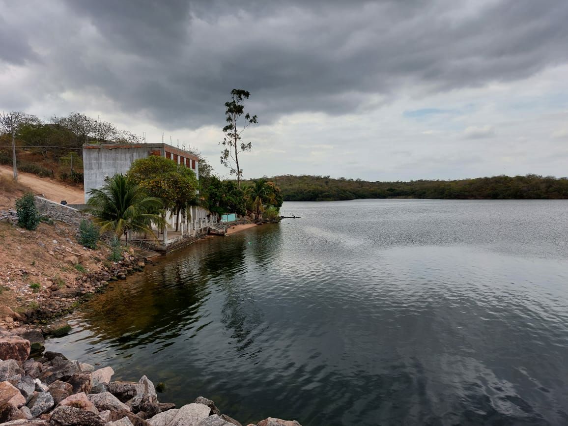 A tranquil lakeside view features a calm, reflective water surface outlined by a rocky shoreline. A building with greenery surrounds it stands in the background, with trees lining the opposite bank. Overcast skies frame the peaceful landscape.