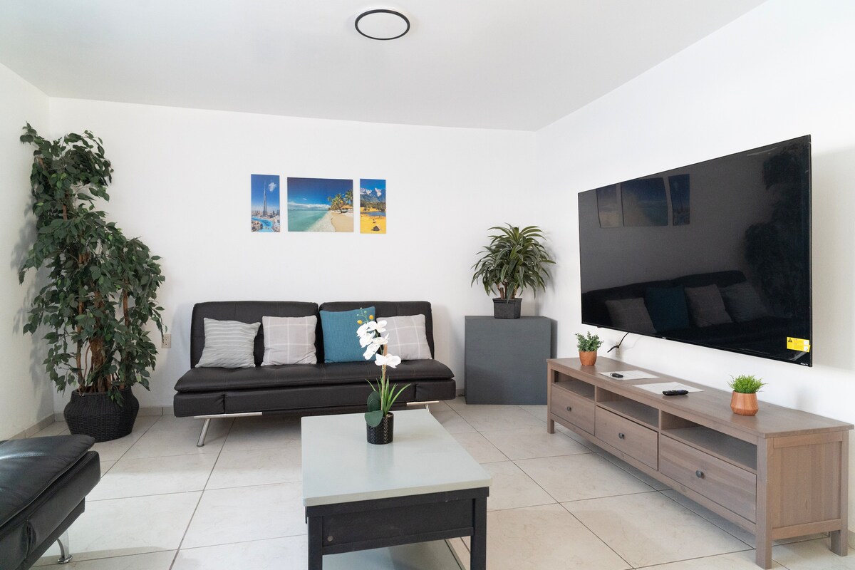 A well-lit living area features a contemporary sofa with cushions and a dark armchair. A large flat-screen TV is mounted on the wall opposite a wooden console table. Plants are placed in the corners, and framed beach prints are displayed above the sofa.
