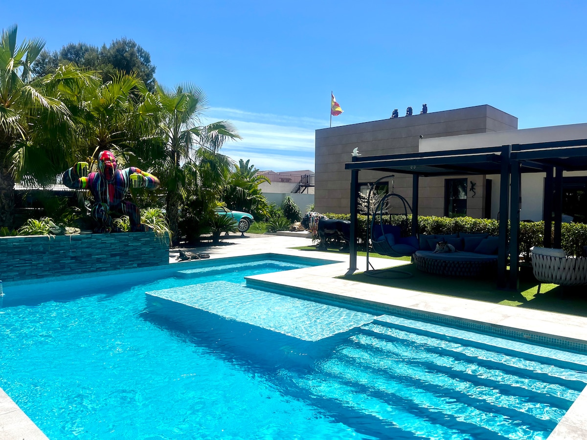 A spacious outdoor area features a sparkling pool surrounded by tropical plants and palm trees. Sun loungers are positioned near the water, and a colorful decorative element adds a playful touch. The modern villa can be seen in the background, offering a glimpse of its design.