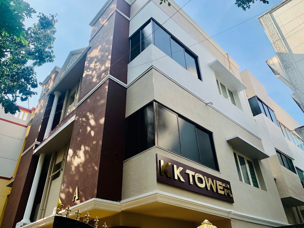 The exterior of the building features a modern design with large windows, allowing for natural light. The name 'KK Tower' is prominently displayed on the façade. Surrounding trees provide greenery, enhancing the welcoming appeal of the entrance.