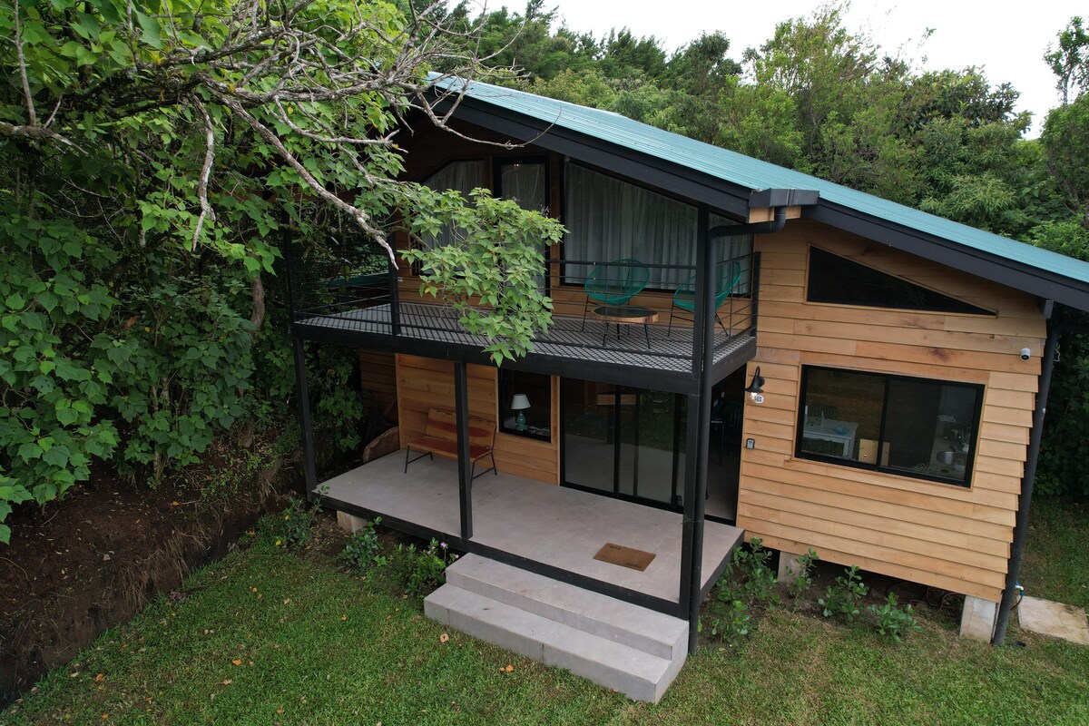 A wooden house featuring a sloped roof is surrounded by lush greenery. A covered deck with two chairs overlooks the grassy area. Large windows provide natural light to the interior, while steps lead up to the entrance, enhancing the connection with the outdoor environment.