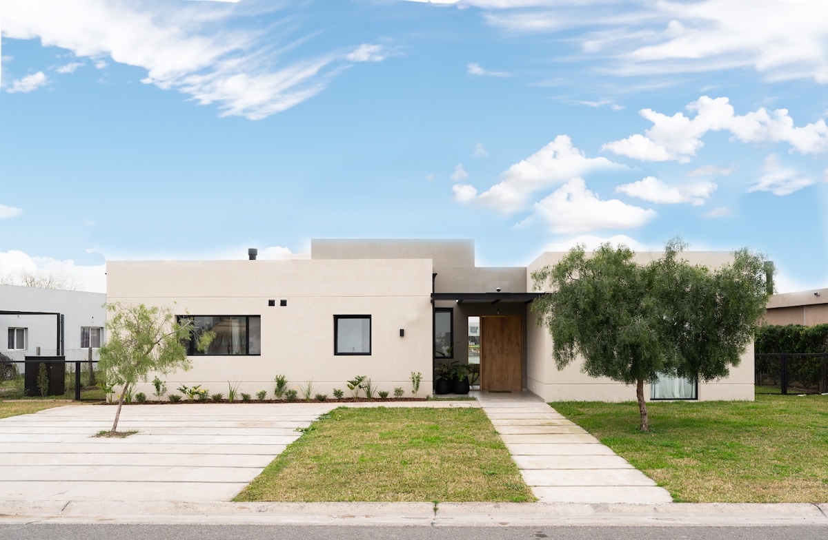 A modern single-story home is presented with a minimalist façade, featuring large windows and a wooden entrance door. The front yard is well-maintained, with green grass and small shrubs. The clear blue sky enhances the property's contemporary appearance.