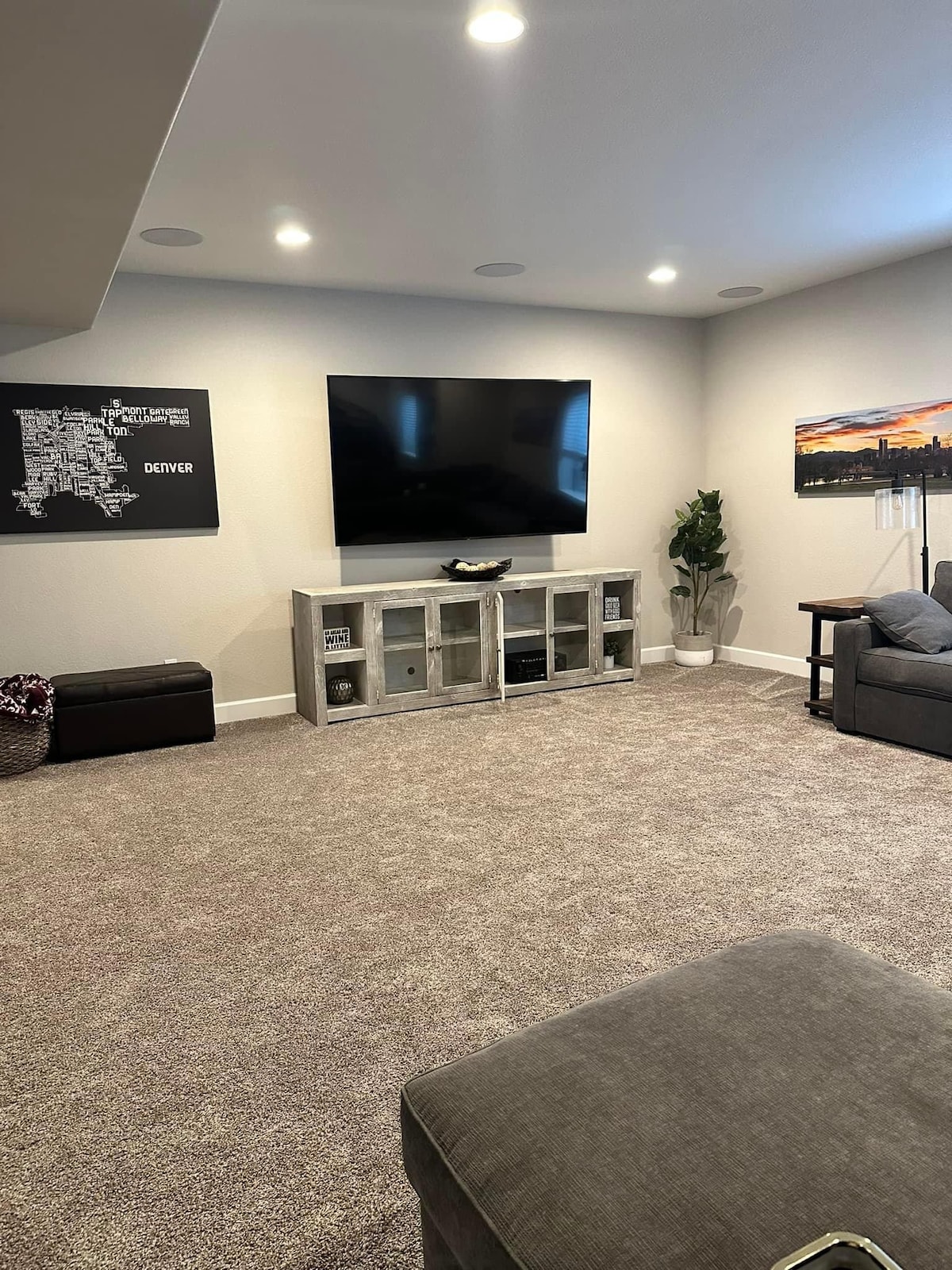 A spacious living area features a large wall-mounted television above a low cabinet with a decorative bowl. A plant adds a touch of greenery next to a window displaying cityscape art. Soft carpeting covers the floor, complemented by a cozy seating arrangement.