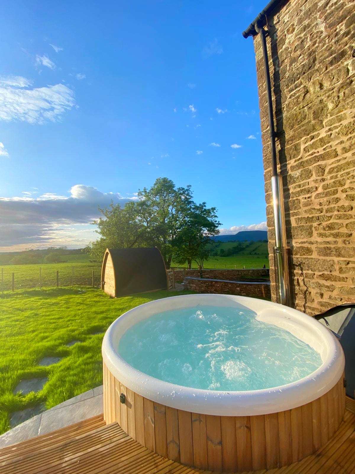A wood-fired hot tub is situated on a wooden deck, surrounded by lush green grass. The expansive landscape features trees and rolling hills under a clear blue sky, creating a serene outdoor setting.