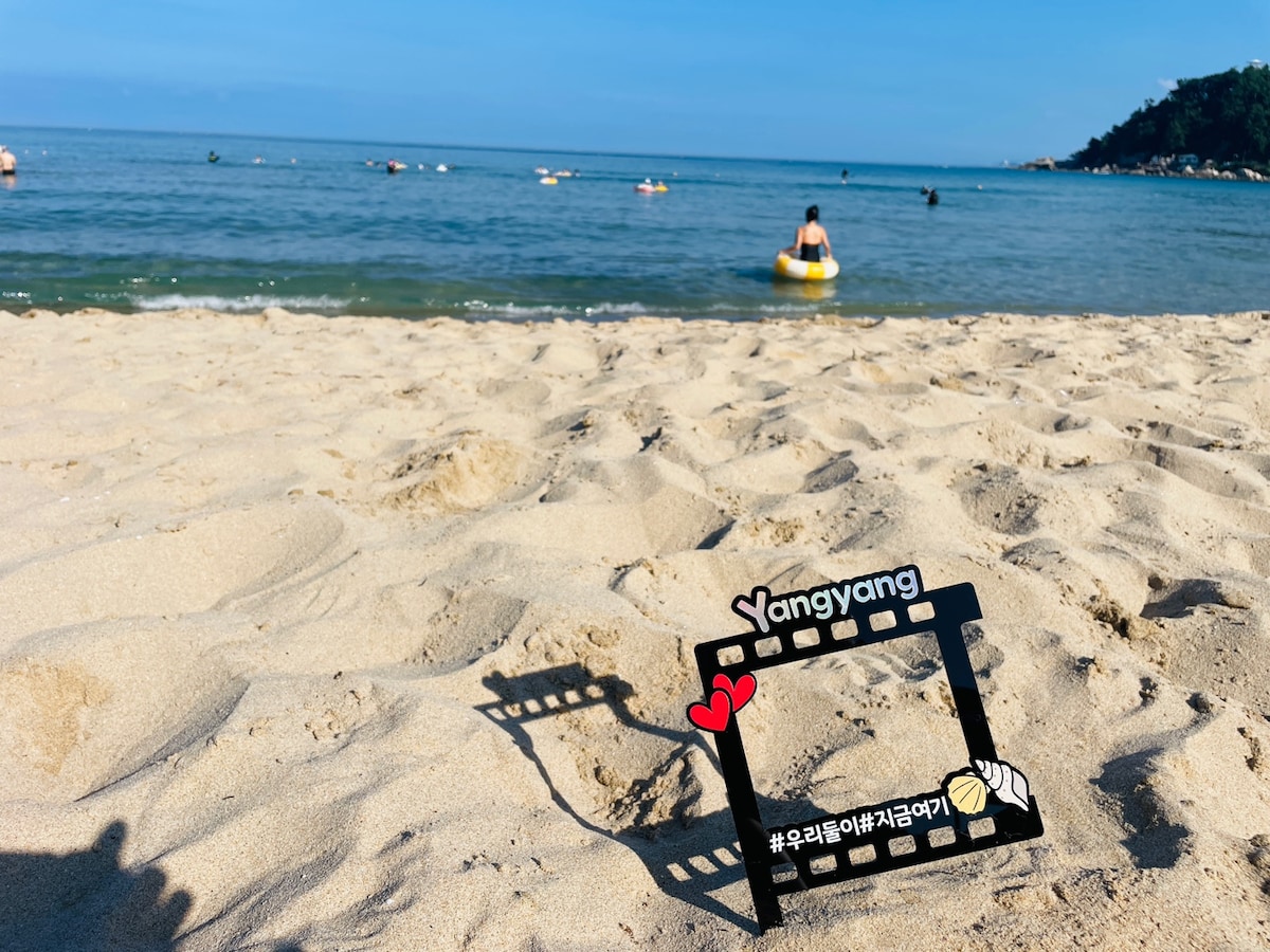 A sandy beach is depicted, with gentle waves lapping at the shore. A colorful film frame prop is positioned in the foreground, featuring the text 'Yangyang' and playful icons. In the distance, beachgoers can be seen enjoying the water on inflatable devices.