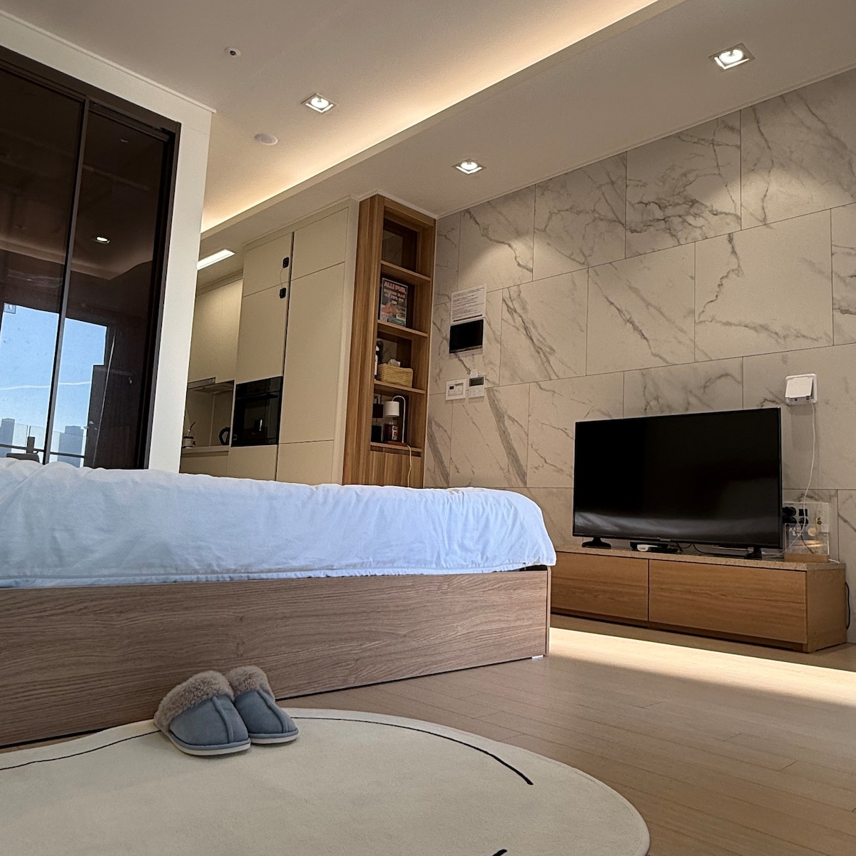 A modern bedroom is featured, showcasing a spacious bed with a light blue duvet. A flat-screen TV is mounted on the wall across from the bed, complemented by a wooden TV stand. Soft slippers are placed on a circular area rug, enhancing the inviting space.