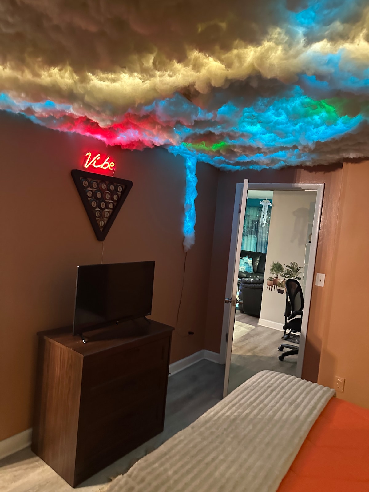 A cozy sleeping area is characterized by a unique cloud-themed ceiling illuminated in various colors. A wooden dresser serves as a base for a television, while a door is visible, leading to additional space. Warm walls contribute to a comfortable ambiance.