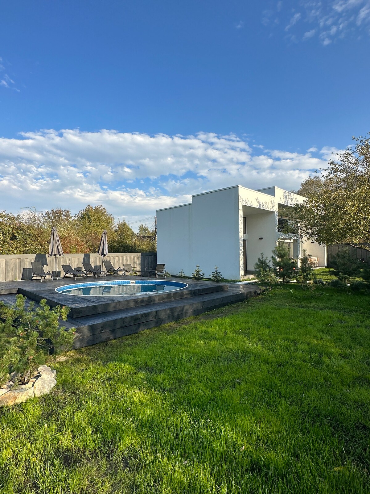 A modern structure is surrounded by lush green grass, featuring a circular hot tub on a raised wooden deck. Several lounge chairs are arranged nearby, set against a backdrop of trees and a clear blue sky with scattered clouds.