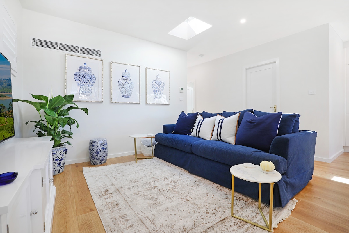 A bright living room is presented, featuring a spacious blue sofa adorned with white and navy cushions. Two framed artworks hang above, complemented by a decorative plant in the corner. Natural light filters through a skylight, enhancing the warm wooden flooring.