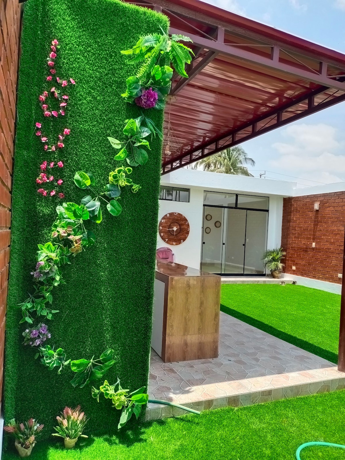 A vibrant green wall adorned with artificial flowers complements a spacious outdoor area. The setting features a well-maintained lawn with a paved pathway, leading to an open indoor space marked by large glass doors and a wooden countertop.