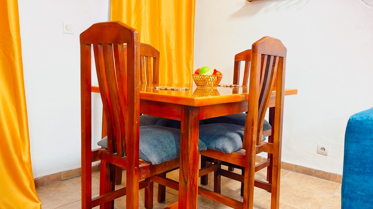 A wooden dining table is surrounded by four tall chairs, featuring blue cushions for comfort. A bowl of colorful fruit sits in the center of the table. Bright yellow curtains frame the window, adding a touch of warmth to the space.