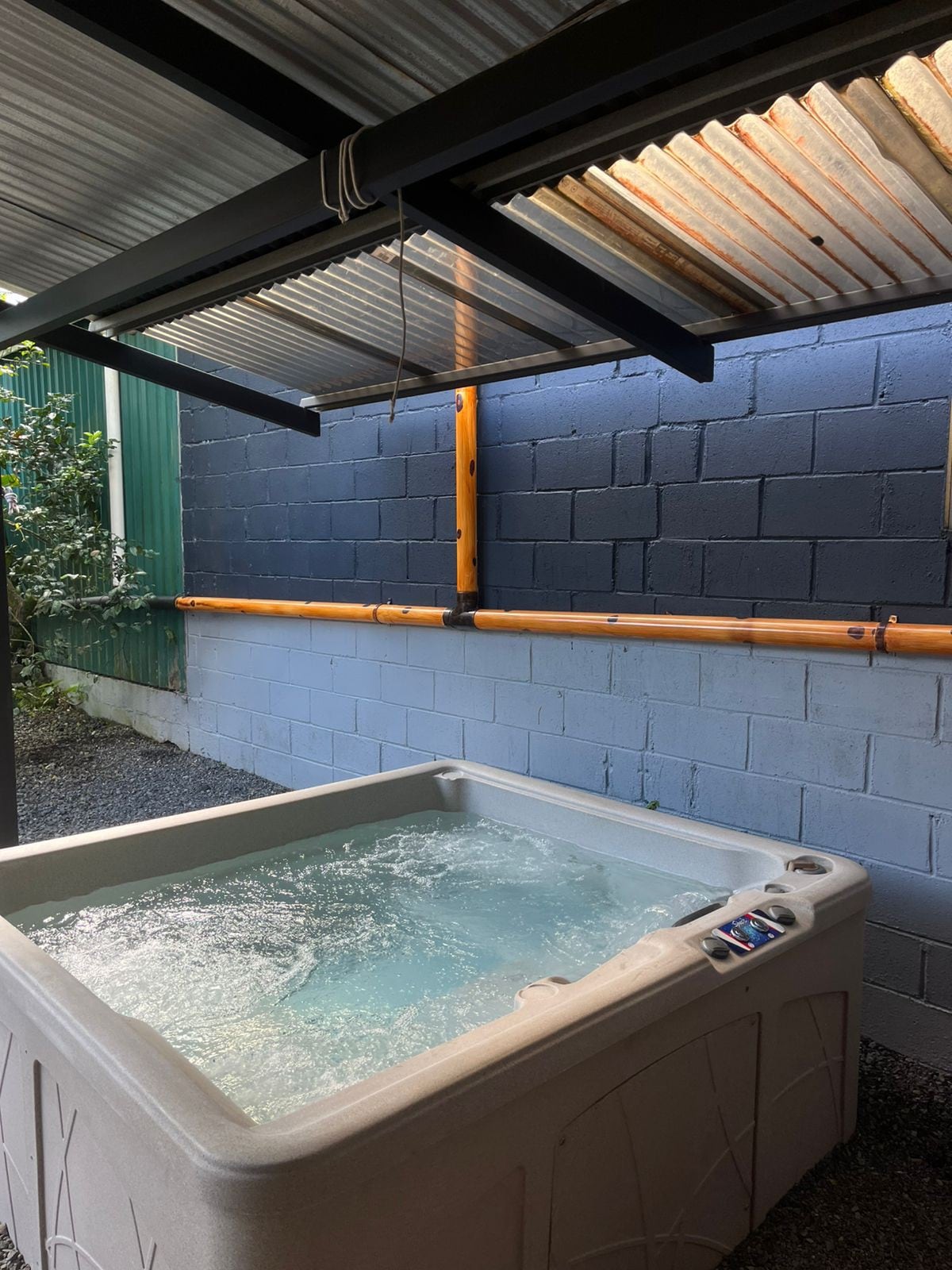 A spacious hot tub is positioned under a covered area, surrounded by blue and green walls. Bordered by stone gravel, the bubbling water invites relaxation in a tranquil setting. The overhead covering provides shade while allowing natural light to filter through.