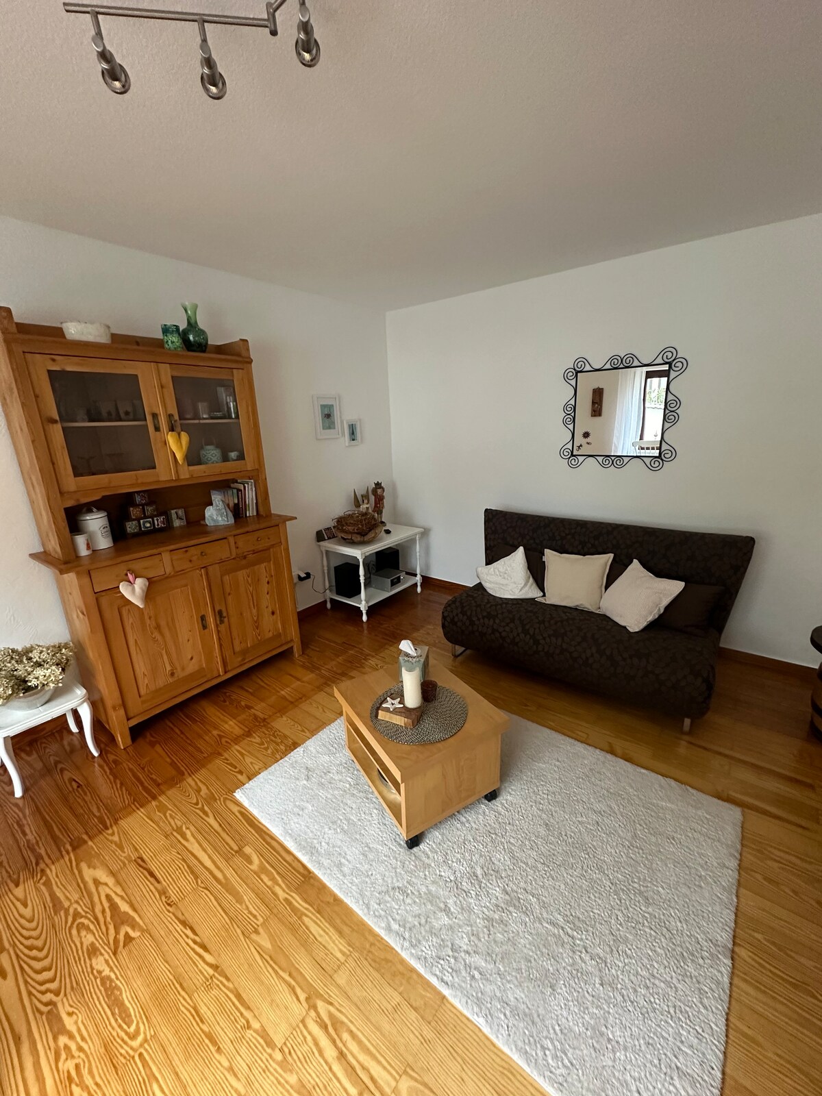 The living area contains a dark brown sofa with white cushions, positioned adjacent to a wooden cabinet filled with decorative items. A small, round wooden table sits on a light-colored area rug, with a candle placed in the center. A mirror reflects the room's light decor.