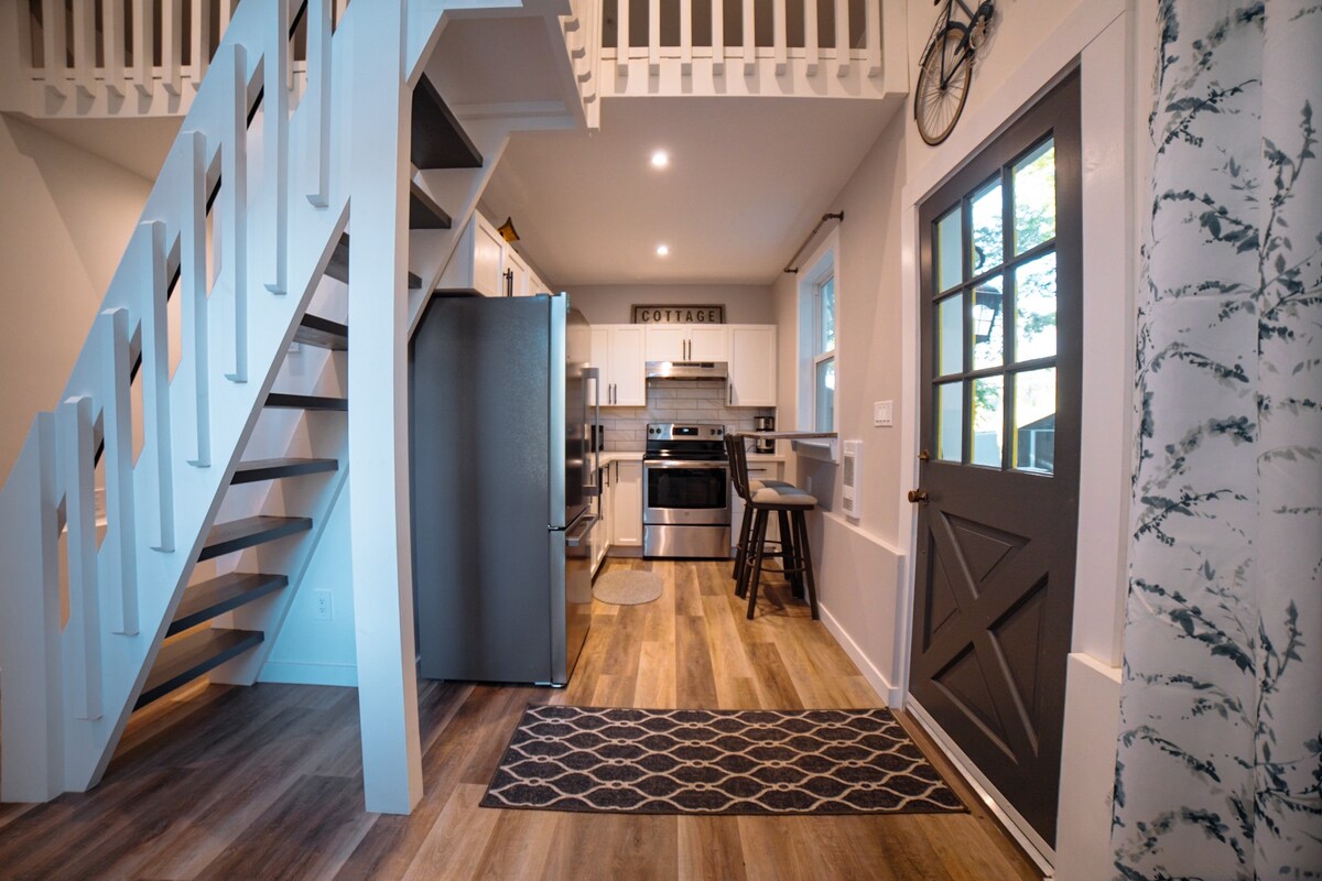 A modern entryway features a functional staircase leading to an elevated loft. Stainless steel appliances are visible in the kitchen, complemented by a patterned area rug and a welcoming door with a window. Natural light brightens the space, enhancing the open design.
