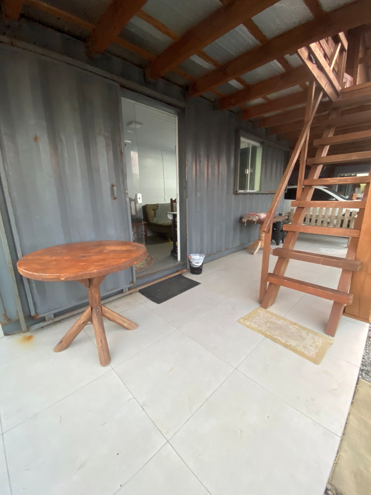 The entrance features a wooden table positioned on a tiled patio, with stairs leading to the upper level. Glass doors provide access to the interior, which is visible through the transparent panels. A small mat lies at the entryway.