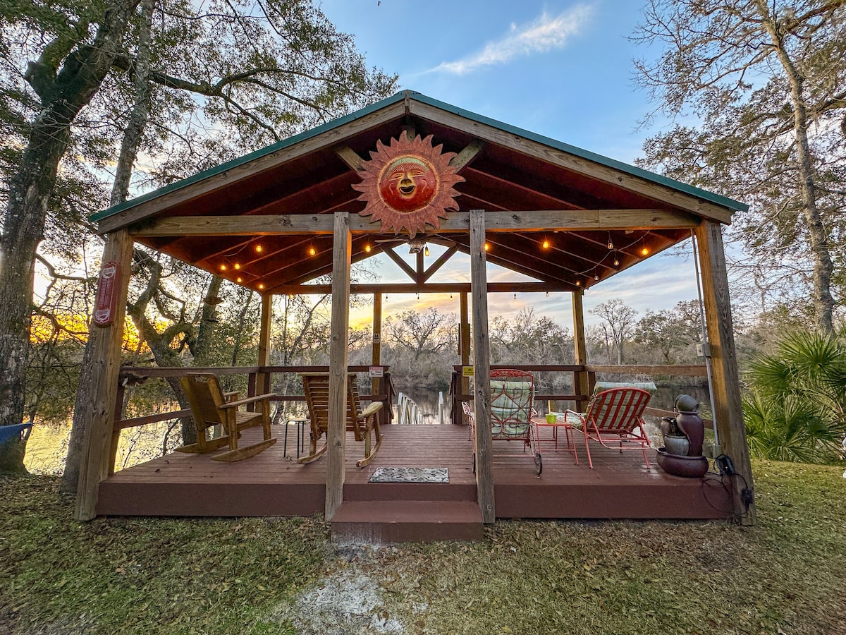 A covered pavilion features a wooden deck with a variety of seating options, including Adirondack chairs and cushioned lounge chairs. Soft lighting casts a warm glow, enhancing the river view framed by trees. A sun motif adorns the pavilion's ceiling, contributing to the inviting ambiance.