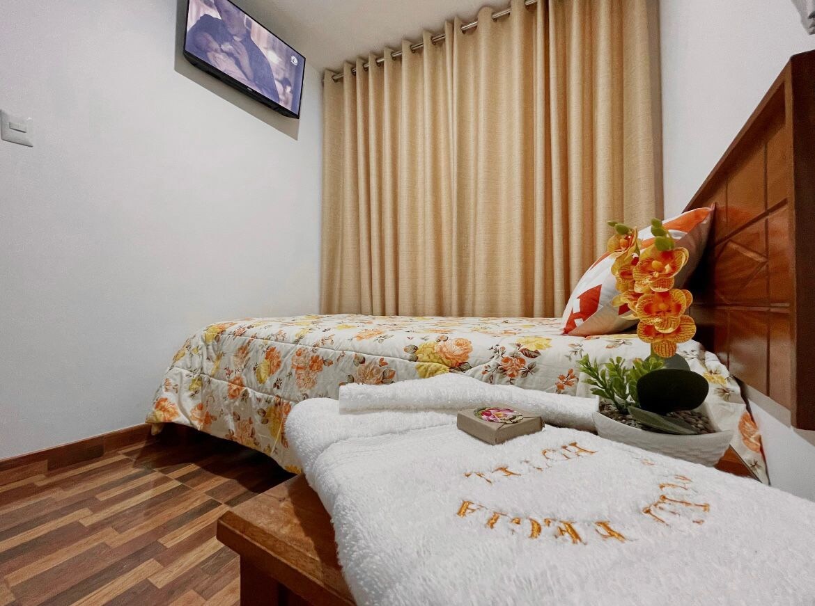 A cozy sleeping area is presented with floral bedding on a single bed. Towels are neatly arranged on a wooden table, accompanied by a small succulent plant and a book. Soft beige curtains frame a window, while a wall-mounted television is visible.