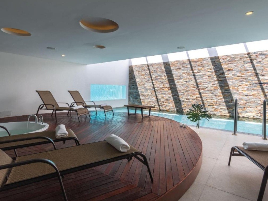 A relaxation area features comfortable loungers arranged around a circular wooden deck. Natural light filters through large windows, highlighting a stone wall and a serene water feature. A small plunge pool sits to one side, complemented by a green plant.