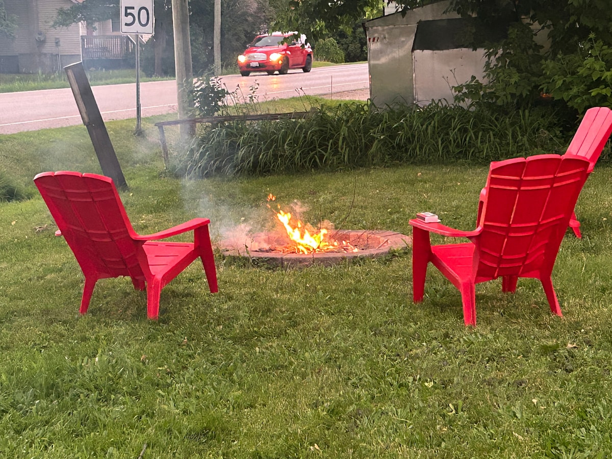 A cozy fire pit area is set on a grassy lawn, featuring two red Adirondack chairs positioned around a crackling fire. The surrounding landscape includes tall grass and a few nearby trees, with a road visible in the background.