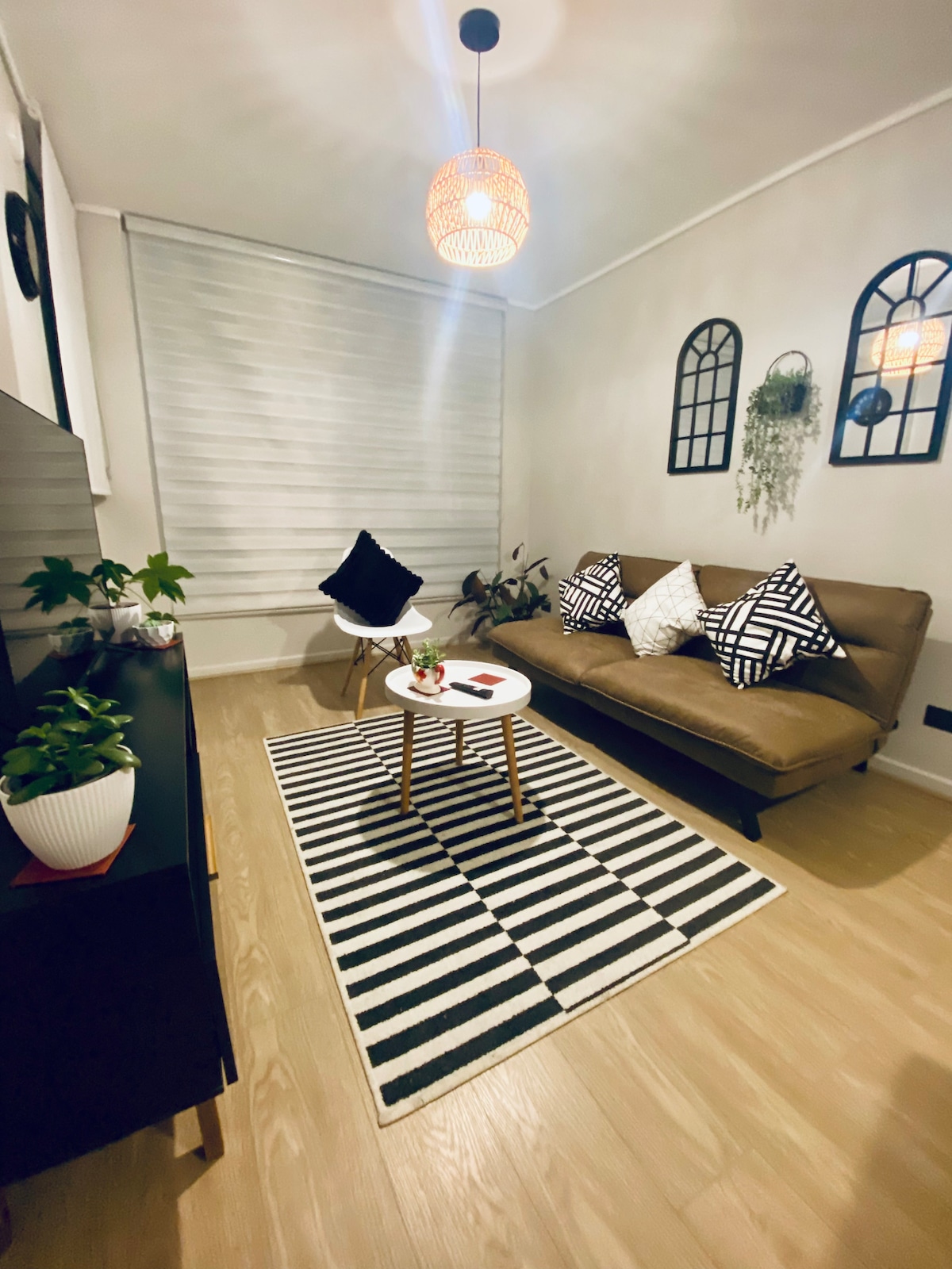 A comfortable living area features a futon, patterned cushions, and a striped area rug. A small round table is set in the center, complemented by decorative plants. Soft lighting is provided by a pendant lamp, and wall decorations include plants and framed mirrors.