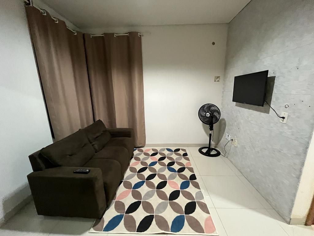 A comfortable seating area features a dark brown sofa positioned against a wall. A modern flat-screen television is mounted above a small console. A patterned area rug with geometric designs complements the space, while a standing fan provides air circulation beside the wall.