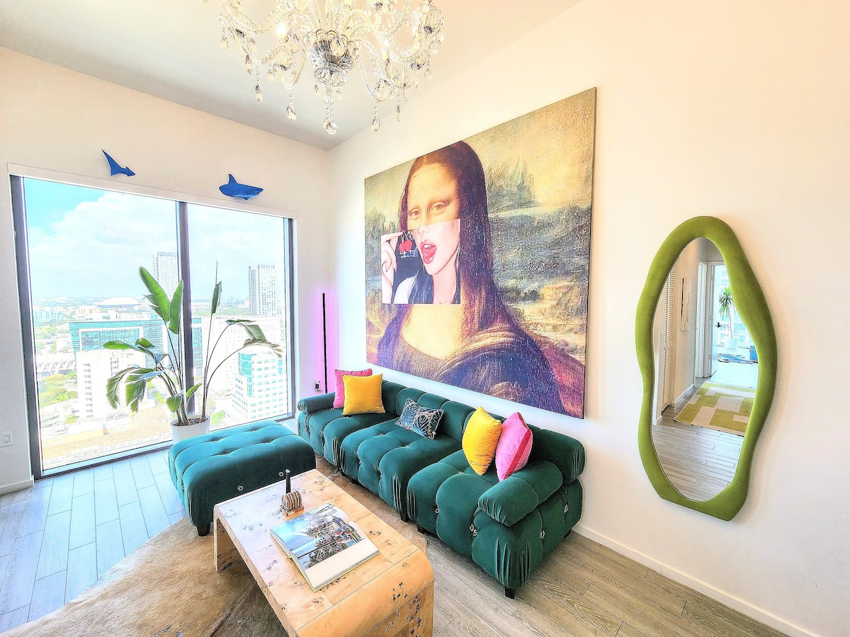A modern living area features a plush green sofa adorned with vibrant pillows. A large, abstract piece of art graces the wall, while a wooden coffee table adds a natural element. Floor-to-ceiling windows illuminate the space, showcasing urban views and a touch of greenery from a nearby plant.