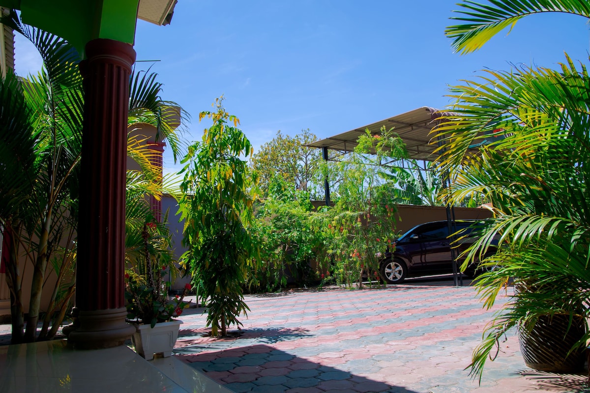 A spacious outdoor parking area is framed by lush greenery. Various plants provide a natural ambiance, while a shaded structure is visible, offering protection for vehicles. A parked car can be seen against the backdrop of a clear blue sky.