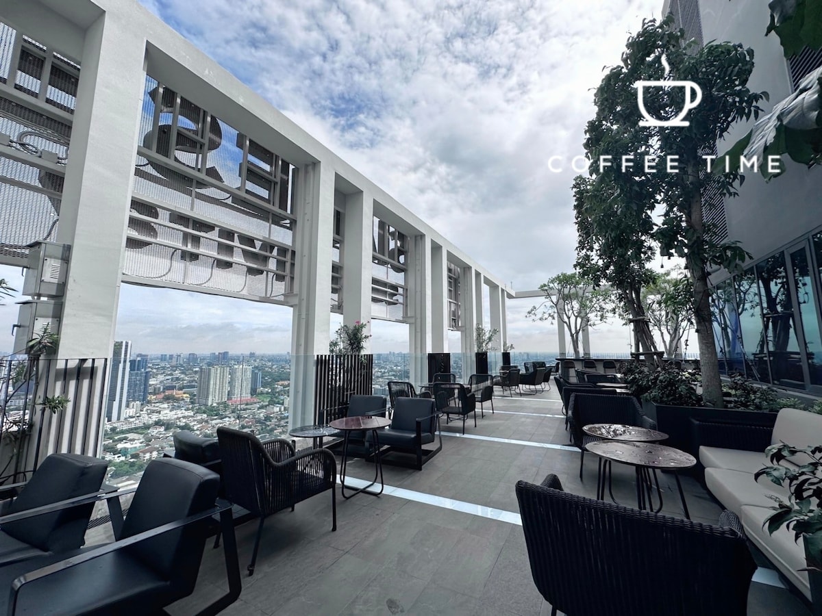 An outdoor terrace is presented with comfortable seating arrangements, featuring black chairs and round tables. Lush greenery is interspersed throughout the space, and expansive views of the city skyline are visible against a partially cloudy sky.