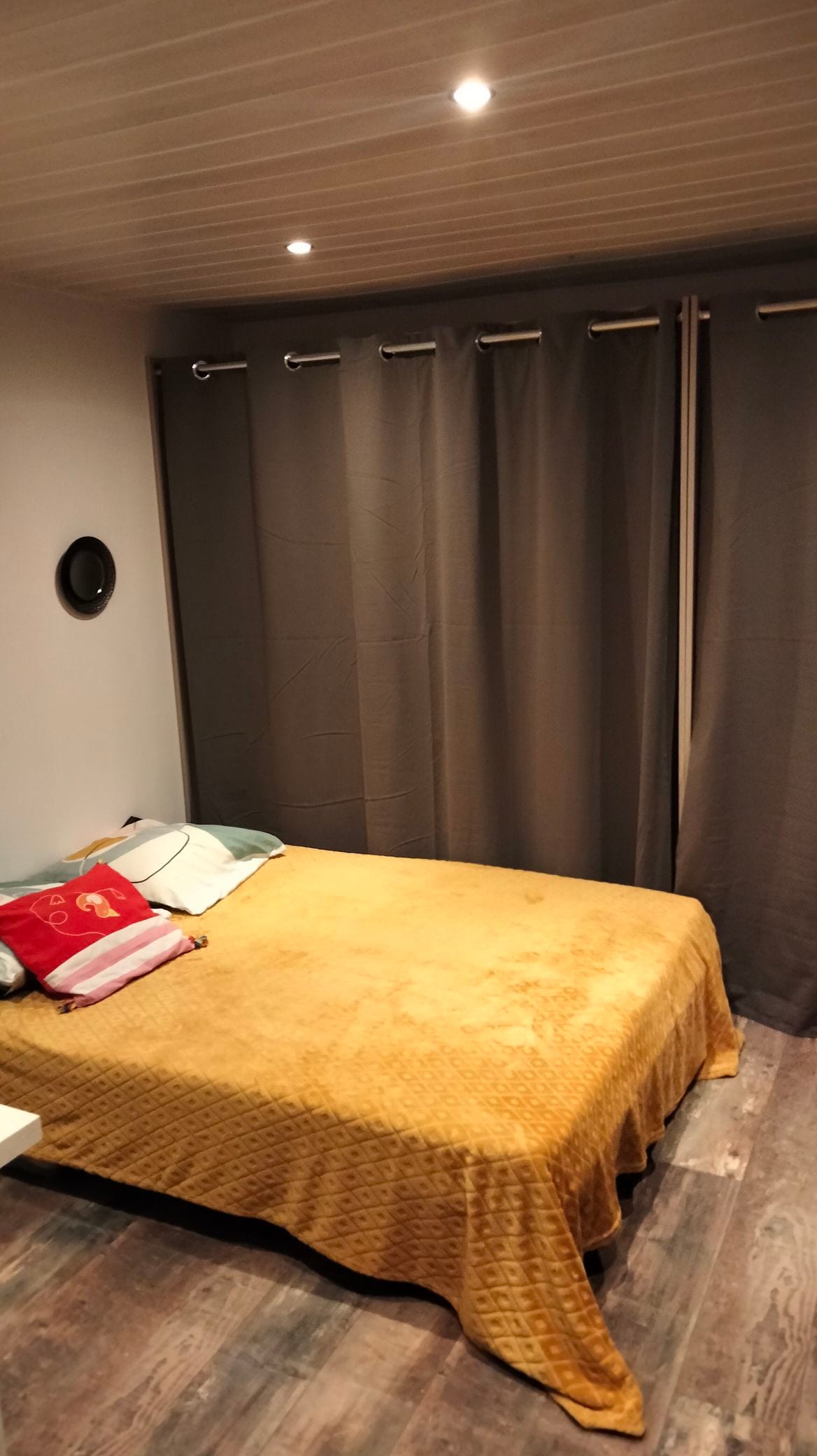 A cozy sleeping area features a spacious bed covered with a golden blanket. Soft pillows are positioned at the head of the bed. Floor-to-ceiling gray curtains frame a window, allowing for privacy and light control. A small round mirror is mounted on the wall.