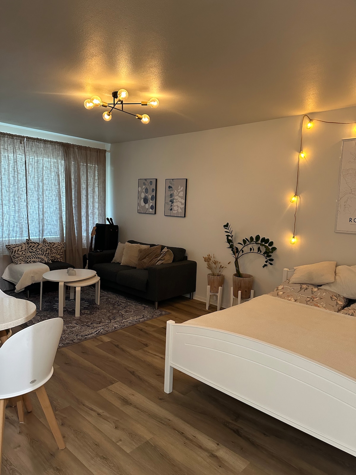 A spacious living area is shown, featuring a comfortable sofa and two round coffee tables. A light-colored bed occupies one corner, while soft lighting illuminates the space. Large windows with sheer curtains allow natural light to enter, complemented by decorative wall art and plants.