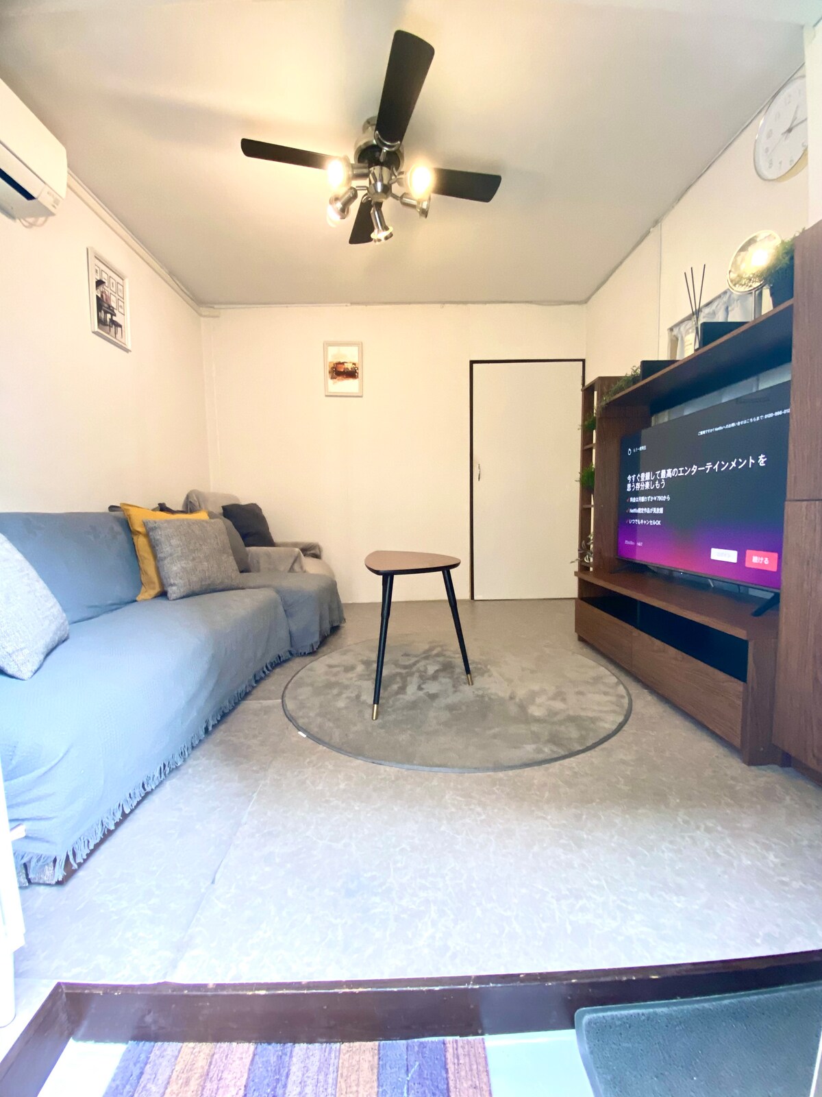 The living area features a cozy seating arrangement with a plush gray sofa adorned with yellow cushions. A small round table sits on a circular rug in front of a large television, providing a space for relaxation and entertainment.