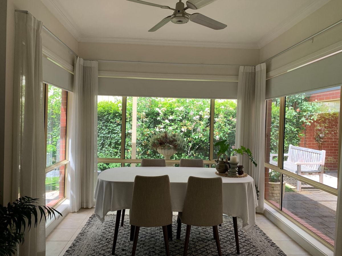 The dining area features a large table surrounded by two upholstered chairs. Full-length windows offer views of the garden, allowing natural light to fill the space. Bright green plants adorn the table, complemented by a neutral rug beneath.