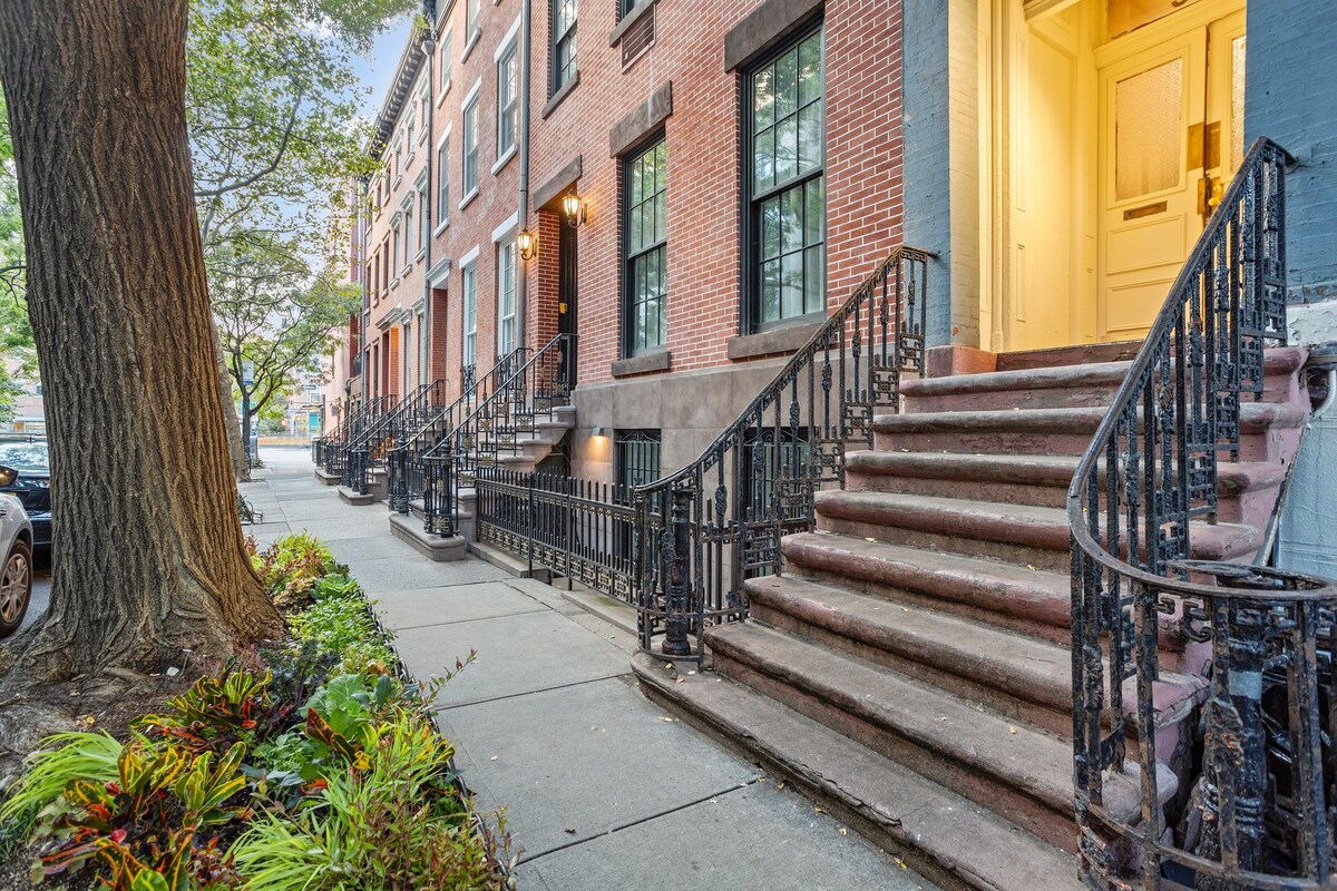 A traditional brownstone building is showcased, featuring a well-maintained exterior with iron railings and steps leading to the entrance. Lush green foliage lines the walkway, enhancing the urban streetscape. The scene is enhanced by gentle evening light filtering through the trees.