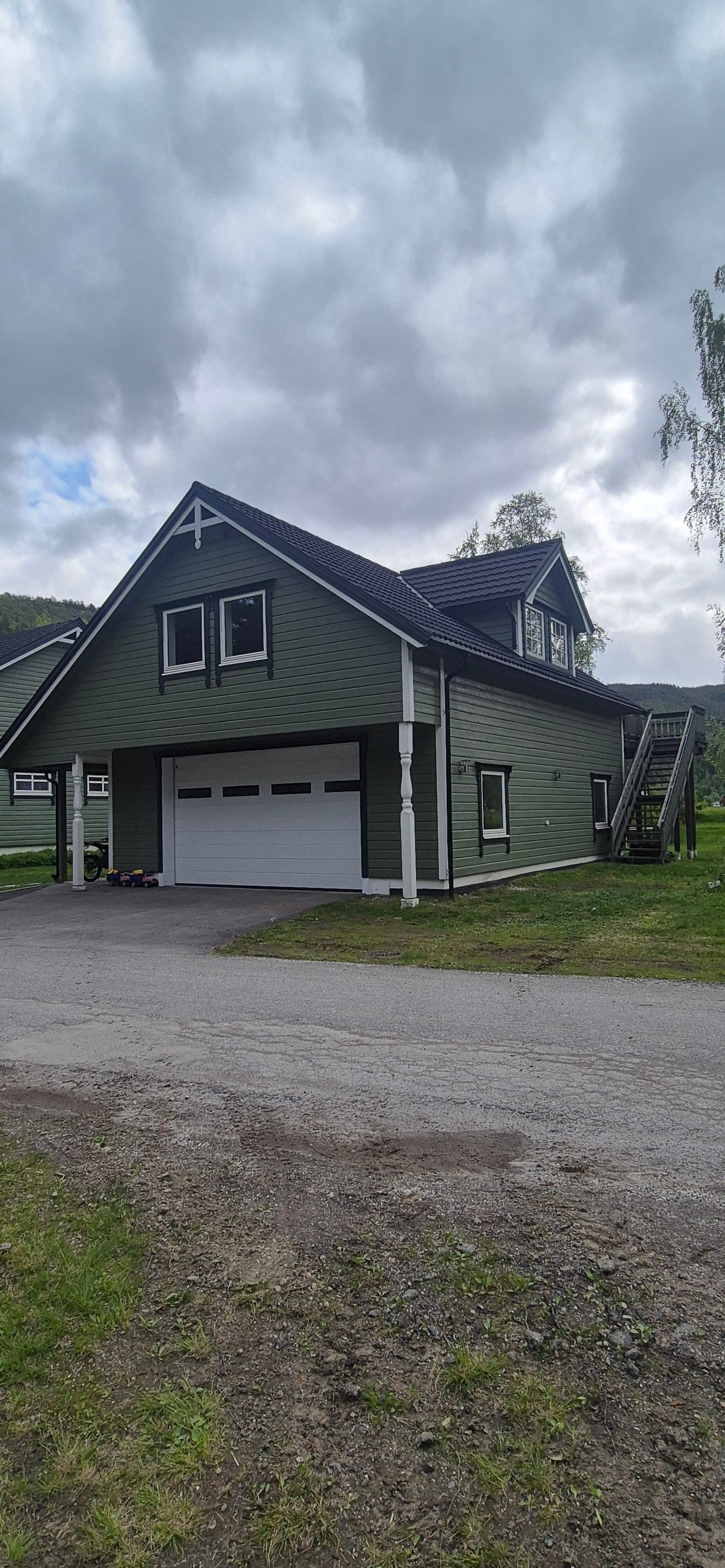 A two-story green building is shown with a steep roof and large garage doors. The entrance features stairs leading to the upper level, which is marked by several windows. Surrounding grassy areas are visible, along with a gravel driveway.