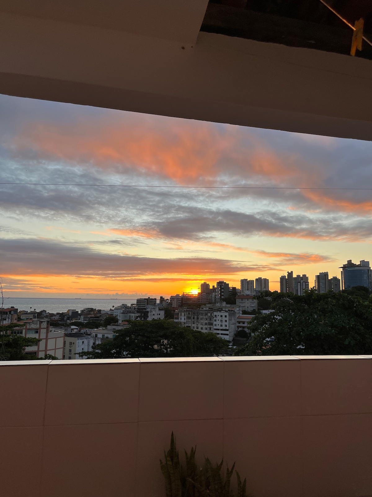 A stunning sunset is visible over the city skyline, with vibrant orange and pink hues reflecting off the clouds. The sea is seen in the distance, framed by the buildings and greenery in the foreground, creating a serene view from the apartment.