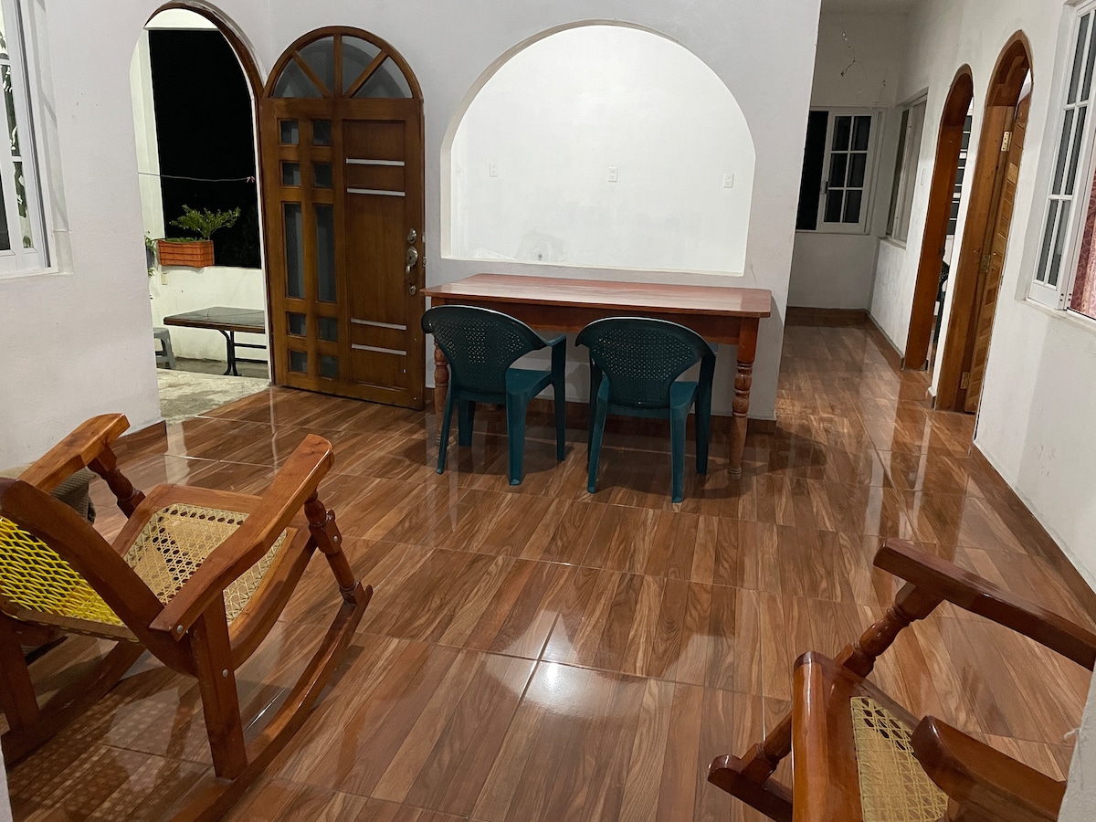 A spacious living area features polished tile flooring, with a wooden dining table positioned centrally. Two green plastic chairs are placed around the table, and two wooden lounge chairs with woven seating are situated nearby. Natural light enters through multiple windows, enhancing the open feel.