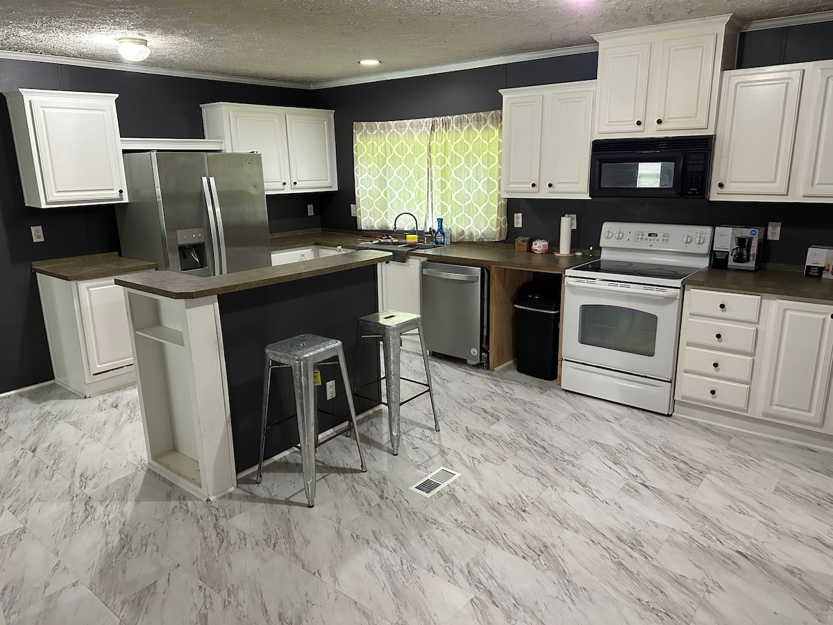 The kitchen area is equipped with white cabinetry and a dark countertop, featuring modern appliances including a refrigerator, stove, and microwave. A window with sheer curtains allows natural light to illuminate the space, complemented by two metal stools at the breakfast bar.