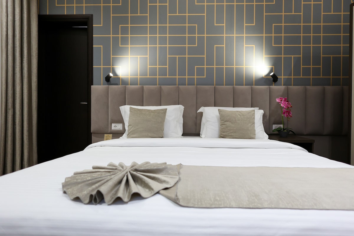 A spacious sleeping area features a comfortable bed with neatly arranged pillows. The headboard is upholstered in a muted tone, complemented by a decorative wall with geometric designs. Soft lighting is provided by wall sconces, and a delicate orchid adds a touch of color.