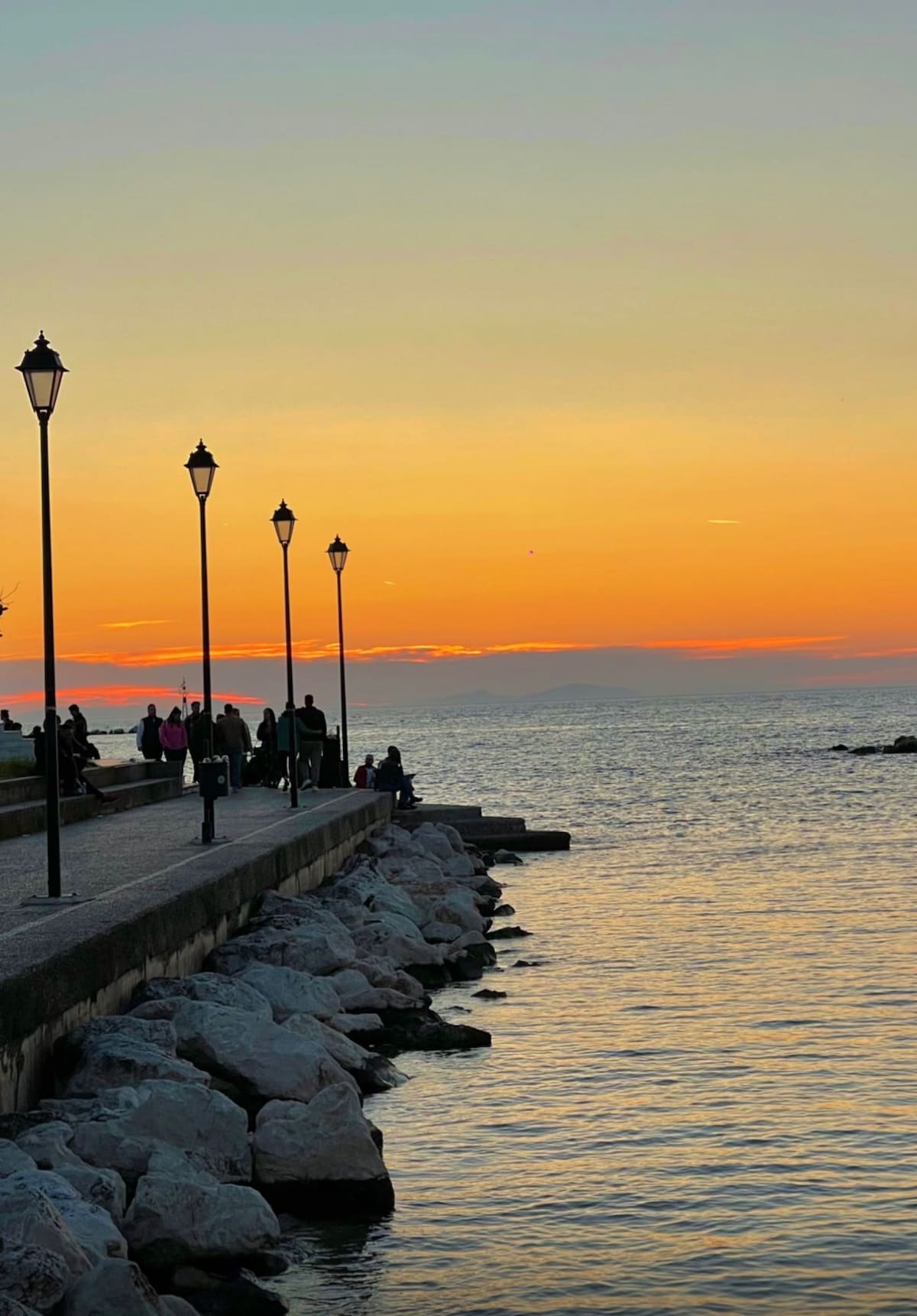 A waterfront promenade is lined with street lamps, casting soft light against the colorful sunset sky. People can be seen strolling along the path, while the gentle waves reflect the warm hues of orange and yellow, creating a serene atmosphere.