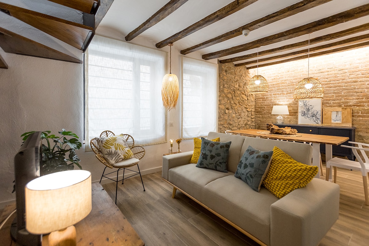A bright living space is revealed with exposed wooden beams and a combination of natural textures. A cozy sofa with patterned cushions faces a wooden dining table. Soft light is emitted from a nearby lamp, enhancing the warm ambiance of the room.