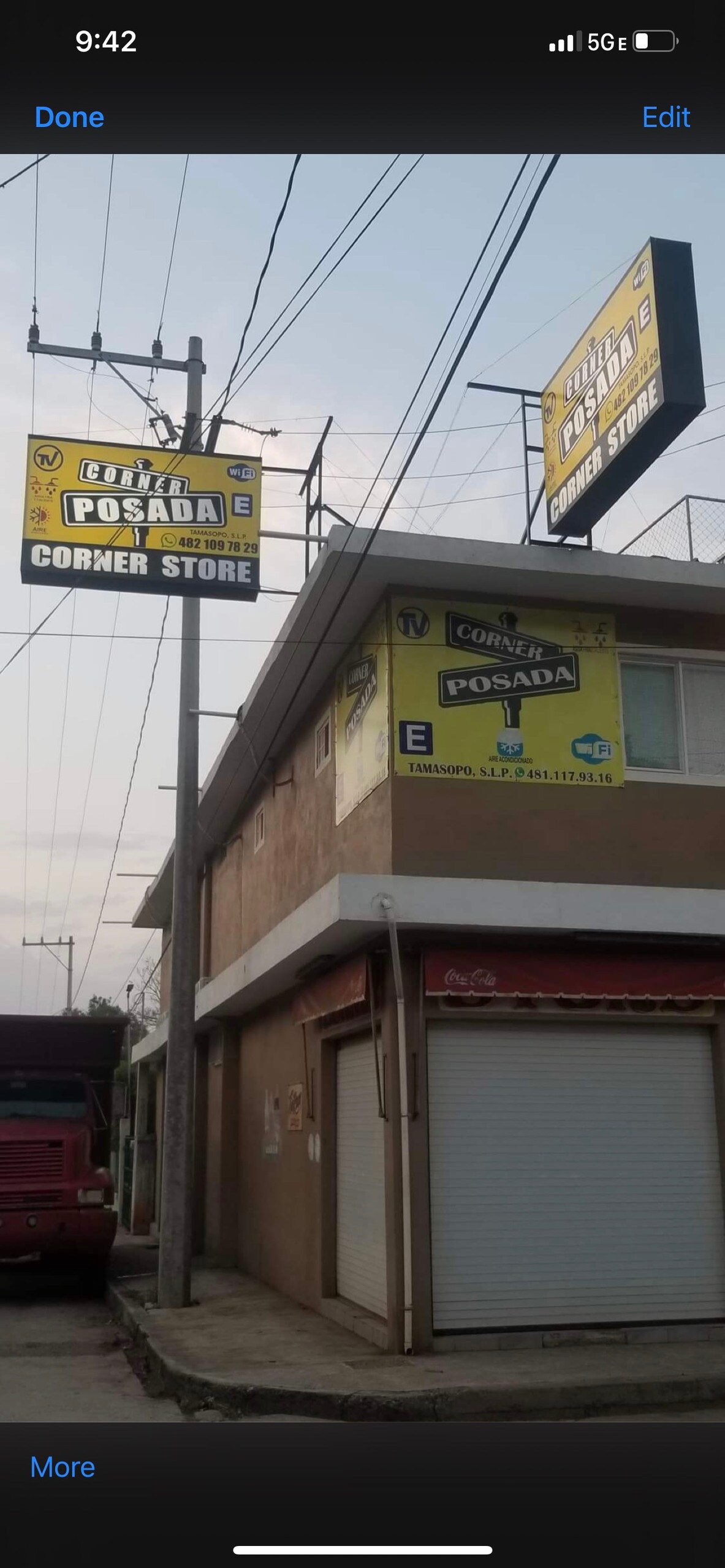 A corner store building is visible, featuring two prominent yellow signs displaying the store's name. The signs are mounted above a light-colored structure with multiple levels. The upper sign identifies the store, while the lower sign includes additional information. The surrounding area has a mix of rooftops and power lines.