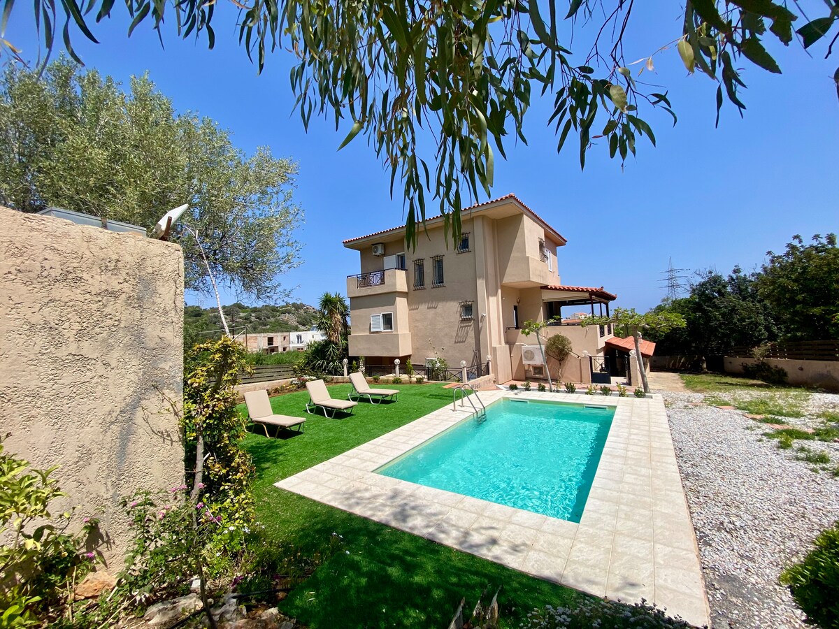 A two-story villa is set amidst lush greenery, featuring a private pool surrounded by pool loungers. The building showcases a traditional architectural style with terracotta roof tiles. The outdoor area includes well-maintained grass, complemented by trees that offer shade.