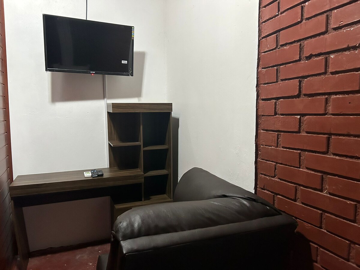 A small living area features a dark leather sofa positioned next to a wooden desk with shelving. A wall-mounted television is visible above the desk, and exposed brick walls add a distinctive touch to the space.