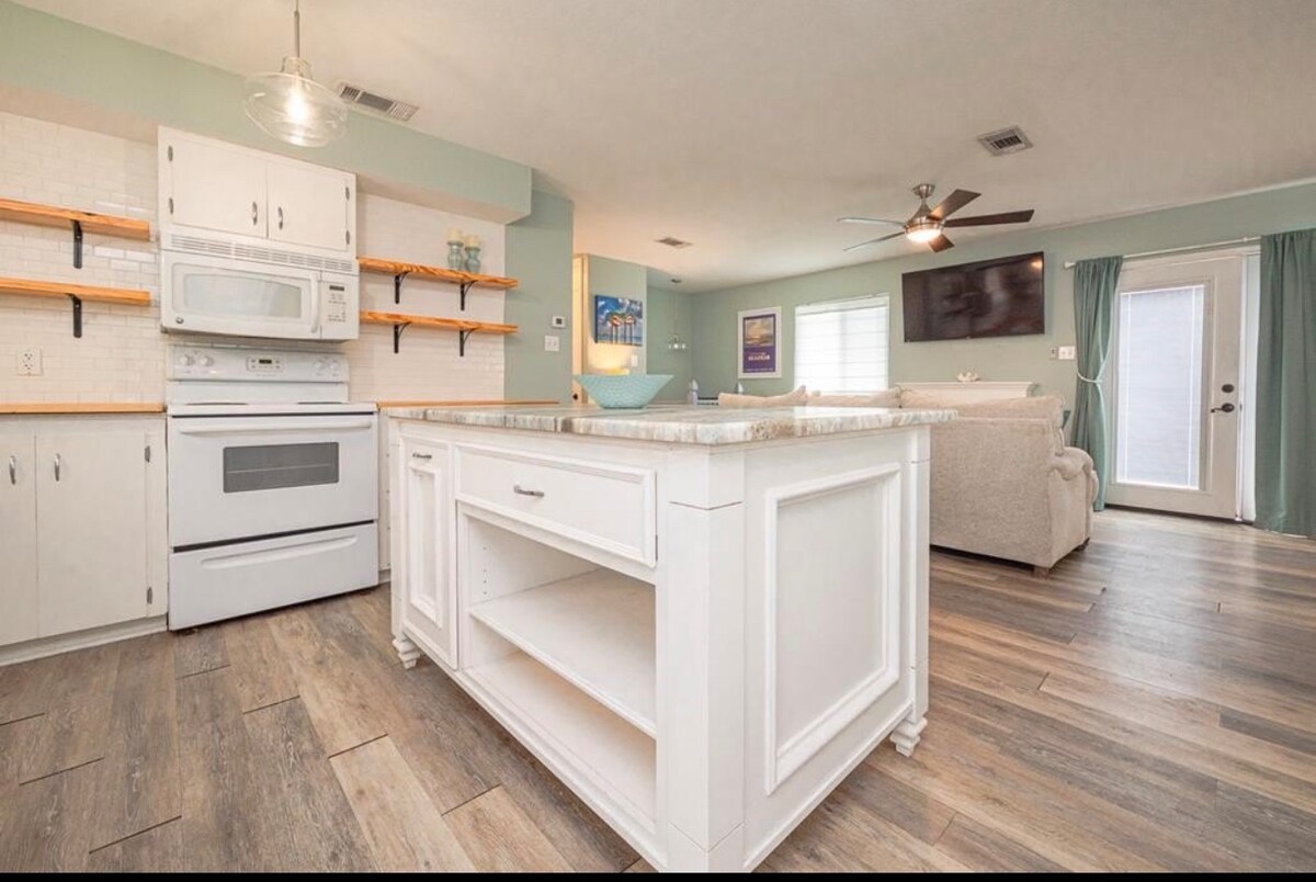 A bright kitchen area features a centrally located island with open shelving. White cabinetry contrasts with light wood flooring. A modern microwave and stove are visible, while a cozy living space with a sofa and wall-mounted television can be seen in the background.