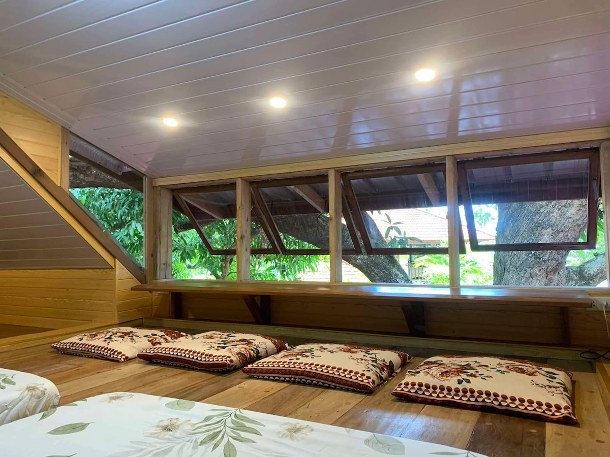 The attic room features wooden flooring and a sloped ceiling lined with recessed lighting. Large windows provide natural light and views of surrounding trees, while several floor cushions are arranged for seating or relaxation.